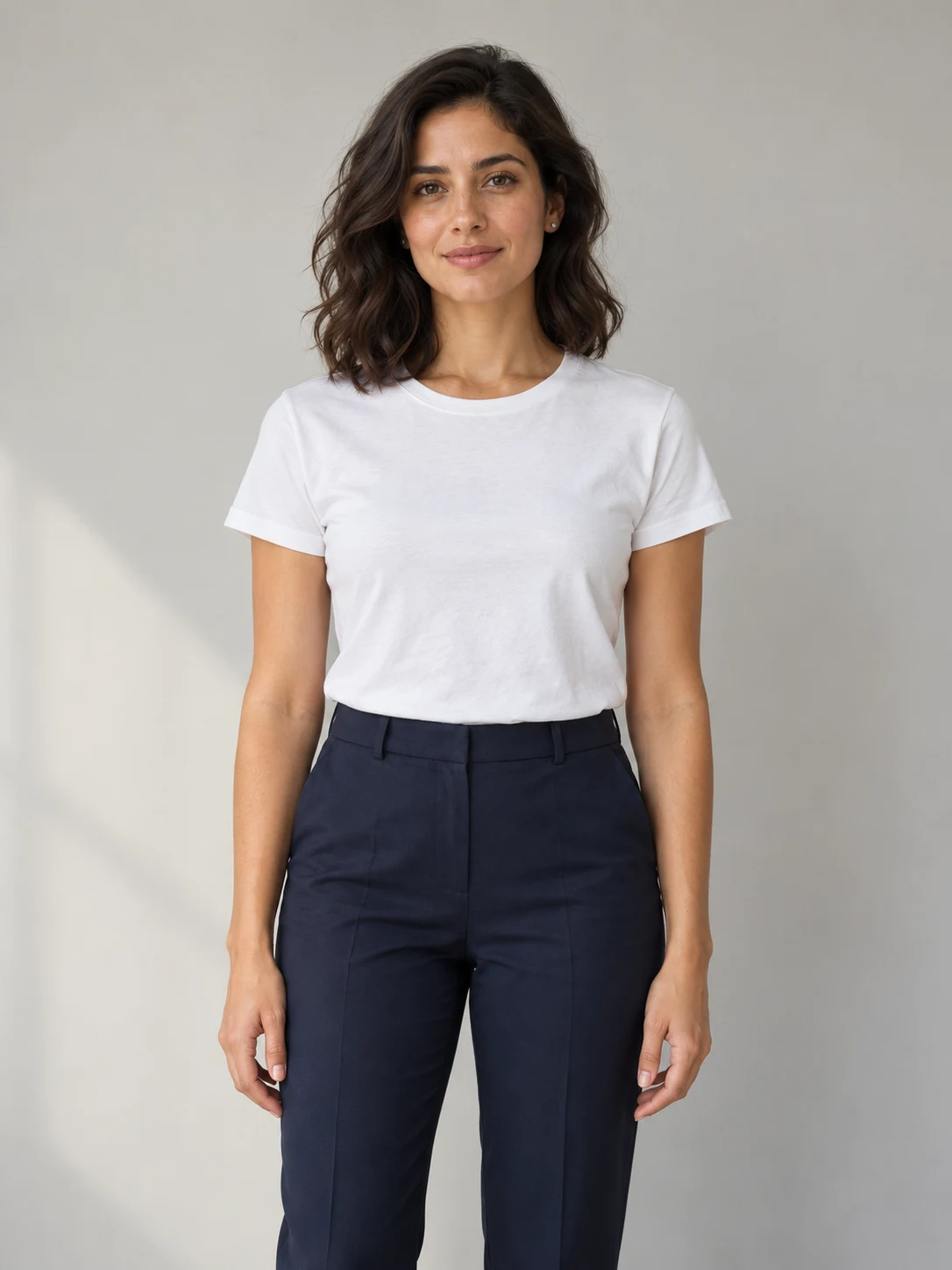 Woman with wavy dark hair, a white T-shirt and navy trousers on a gray backdrop